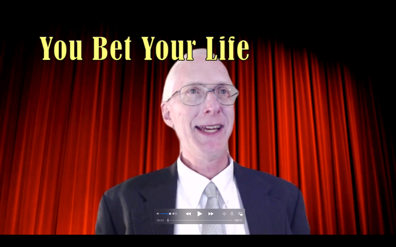 A screen grab from a video, with Dan in a suit and tie apparently standing in front of a red velvet stage curtain, with a title overhead saying You Bet Your Life.
