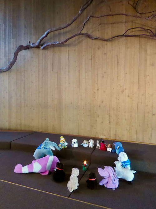 A circle of stuffed animals sitting around a lighted chalice, in a worship space
