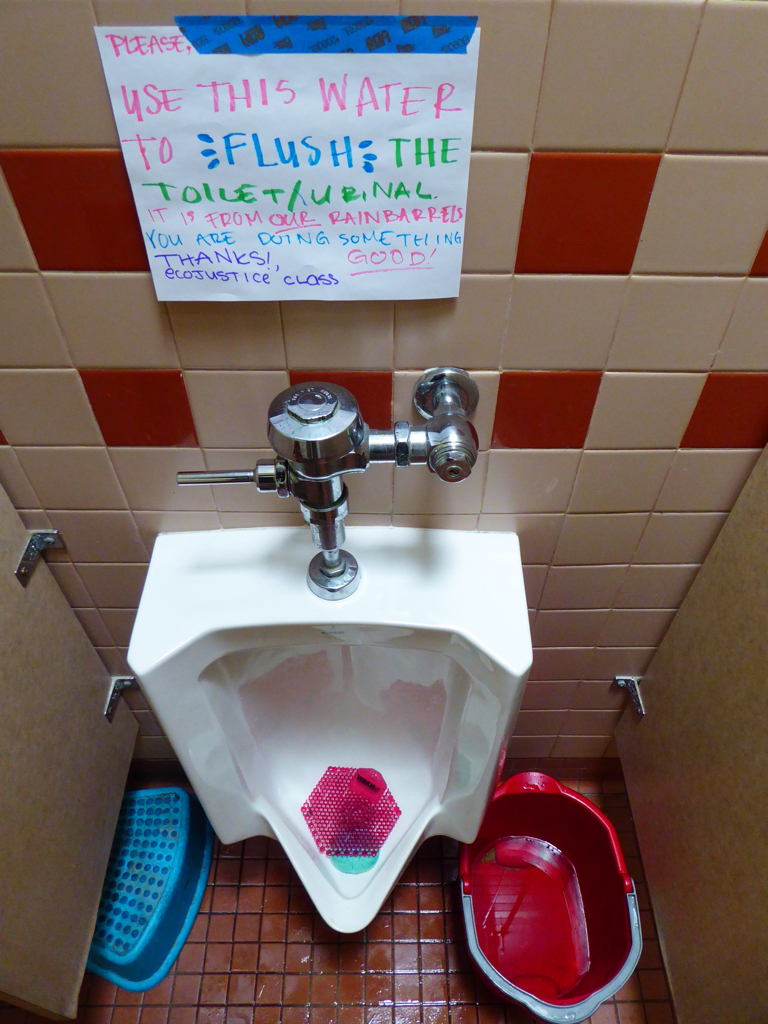 Photo showing a urinal at UUCPA with a partially empty bucket of rainwater next to it, and a sign saying to use the rainwater to flush the urinal