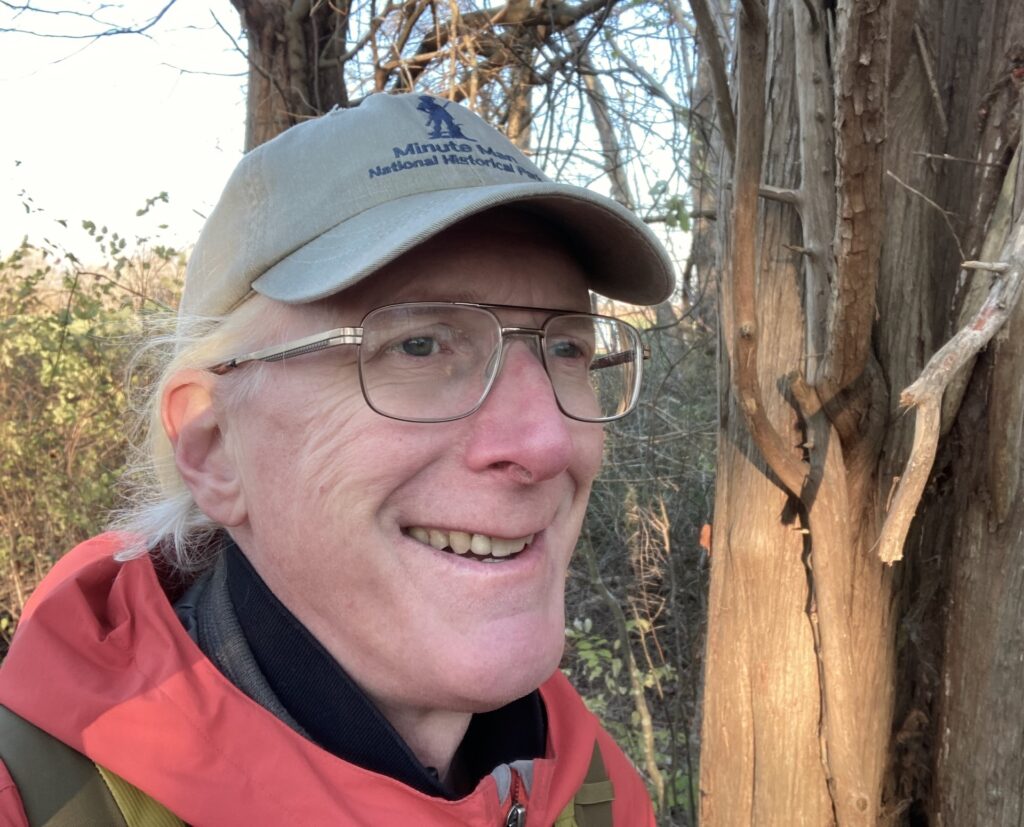 Headshot of Dan standing outdoor in front of a tree trunk.