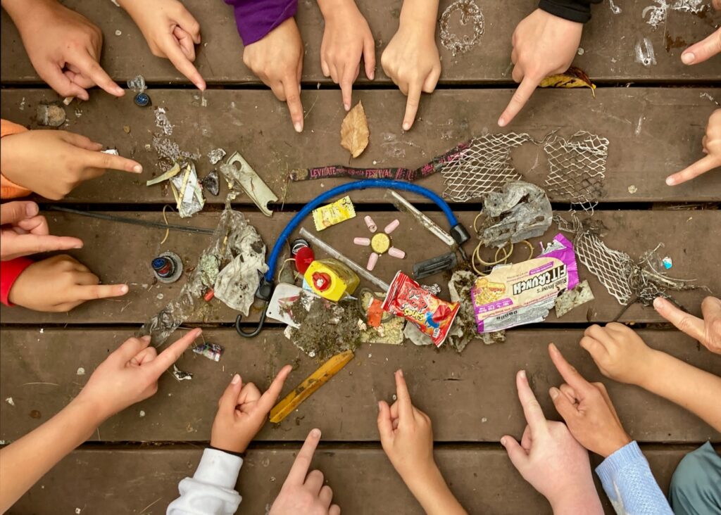 A collection of trash on a picnic table, with hands of children pointing at it.