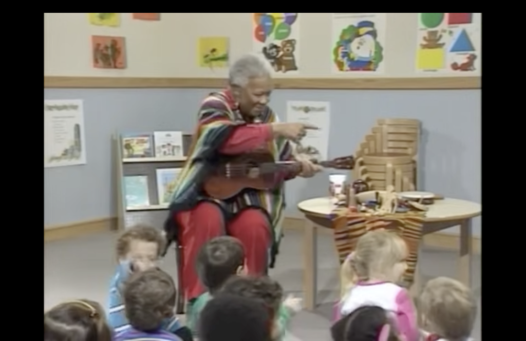 Woman seated on a low chair holding a ukulele, with a group of young children in front of her.