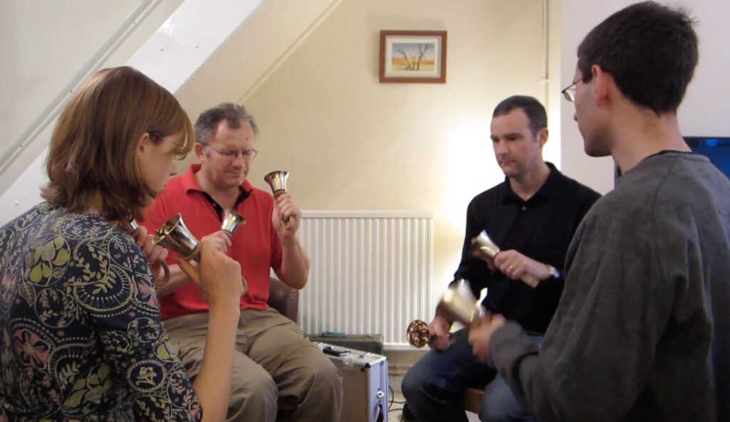 Four people sitting in a circle, each ringing two handbells.