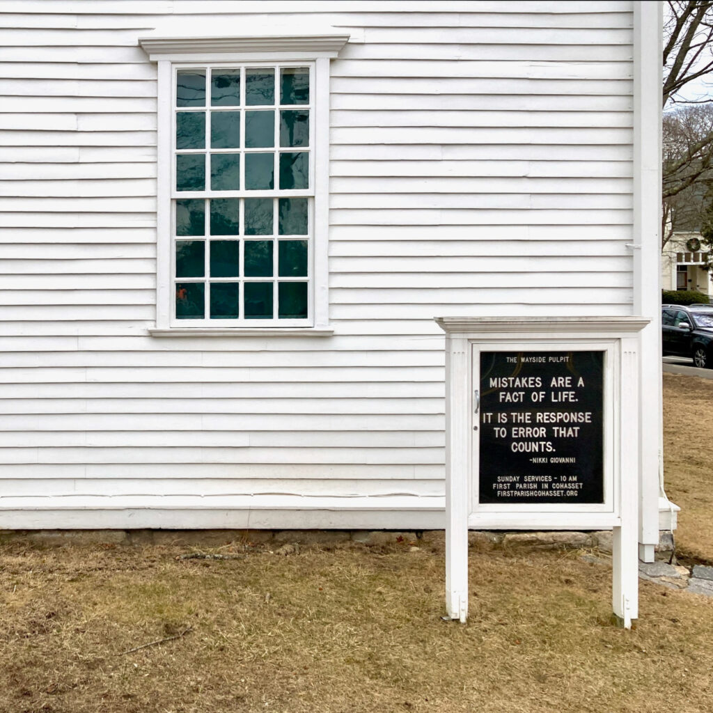 A sign in front of the corner of a New England clapboard meetinghouse.